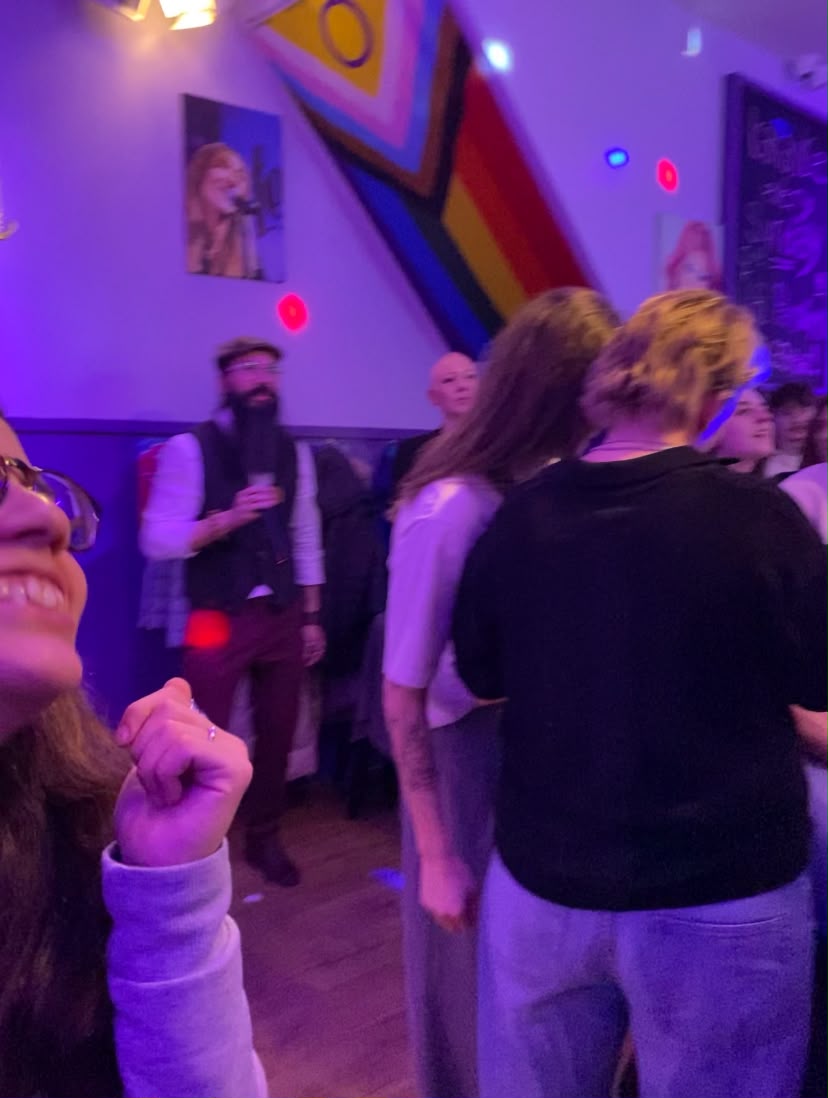 Noite num bar queer com a bandeira Progress Pride na parede