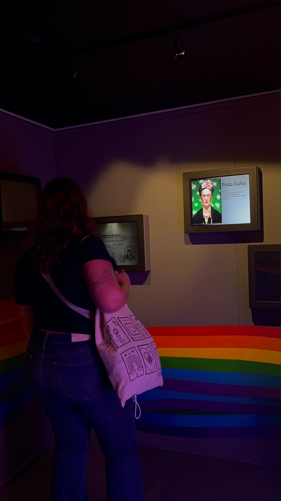 Visitante com uma tote bag a olhar para um ecrã sobre Frida Kahlo, numa sala de museu com um arco-íris na parede