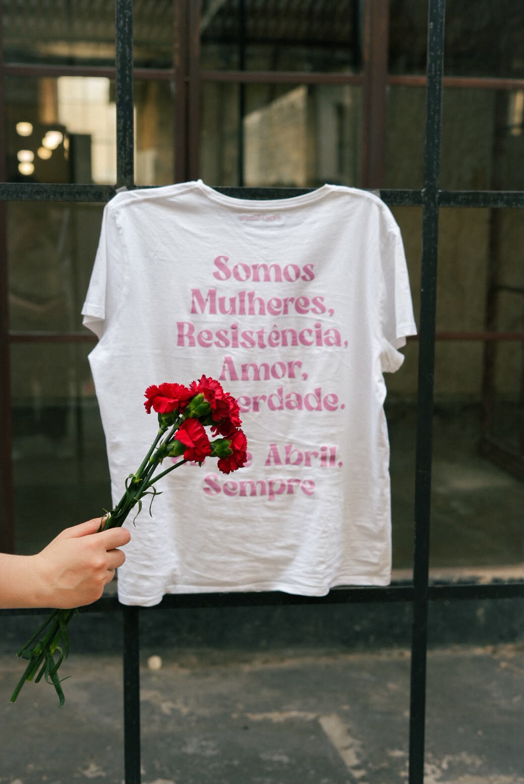 T-shirt branca com a frase 'Somos Mulheres, Resistência, Amor, Liberdade. 25 de Abril, Sempre' e cravos vermelhos