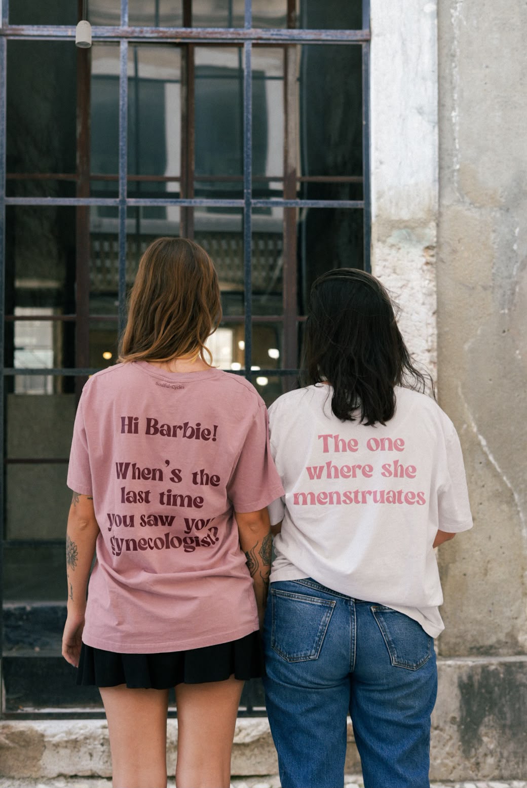Duas pessoas de costas com t-shirts com as frases 'Hi Barbie! When's the last time you saw your gynecologist?' e 'The one where she menstruates'