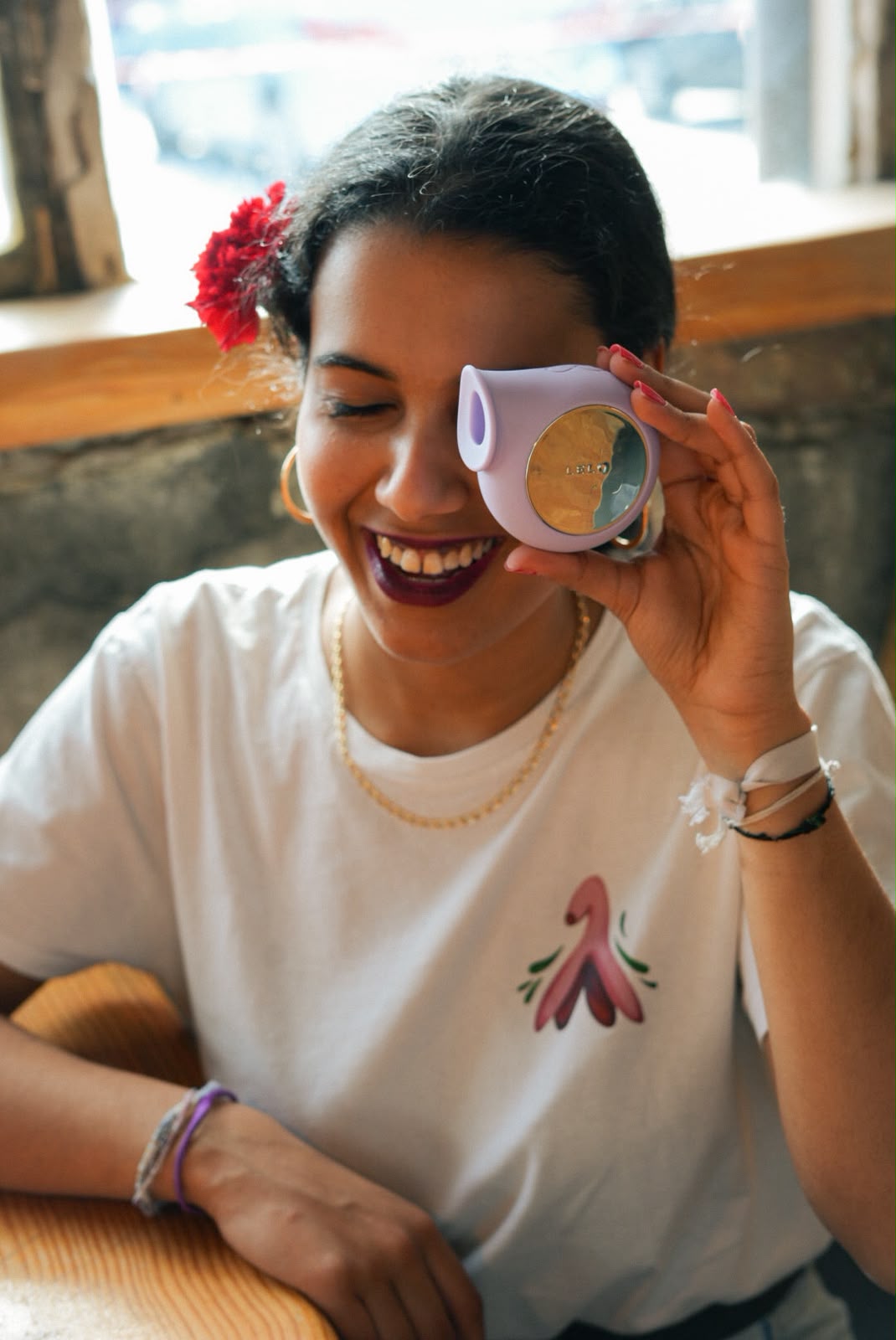 Mulher a rir com uma t-shirt branca com a ilustração de um clítoris, a segurar um brinquedo lilás à frente de um olho