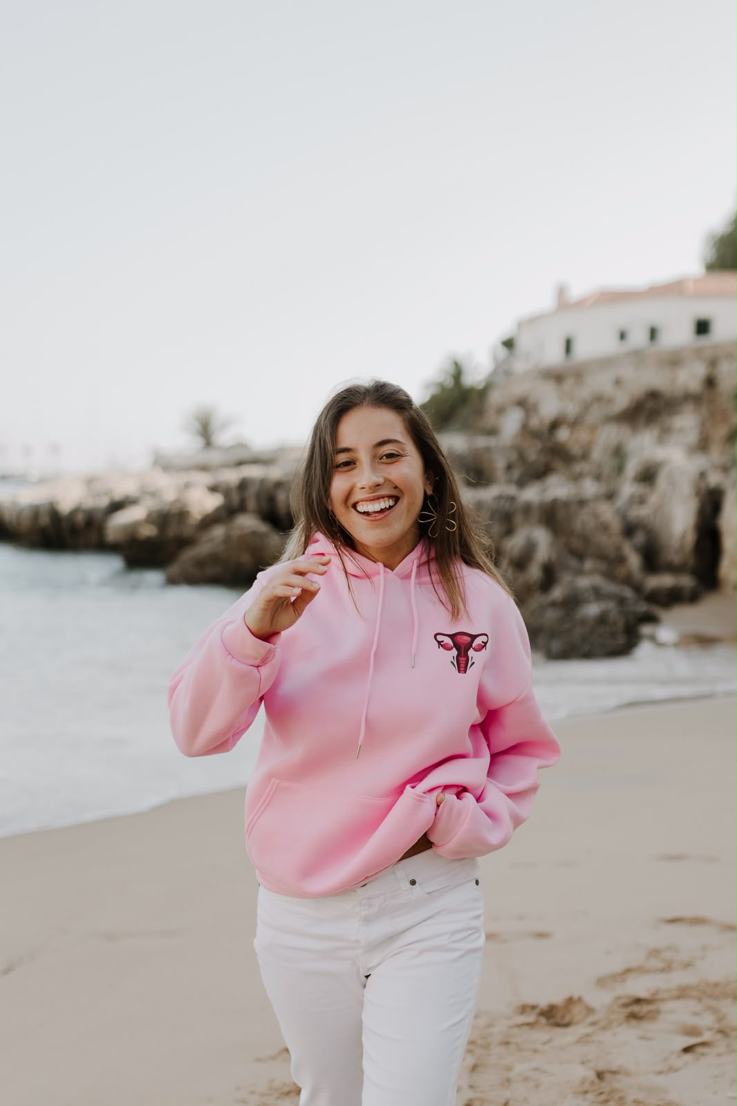 Mulher a rir na praia com uma hoodie cor-de-rosa com a ilustração de um útero