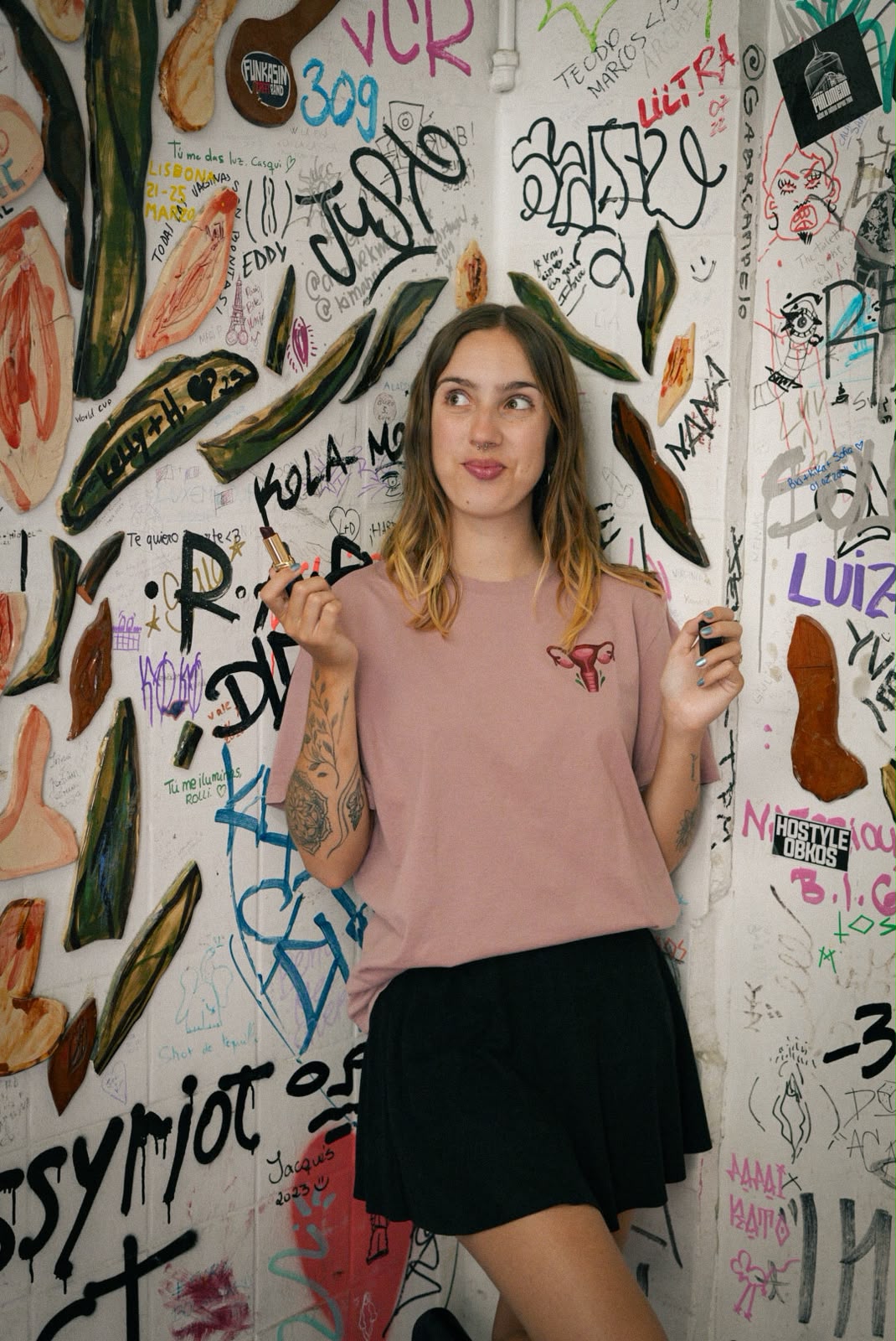 Mulher com uma t-shirt cor-de-rosa com a ilustração de um útero, em frente a uma parede com graffiti