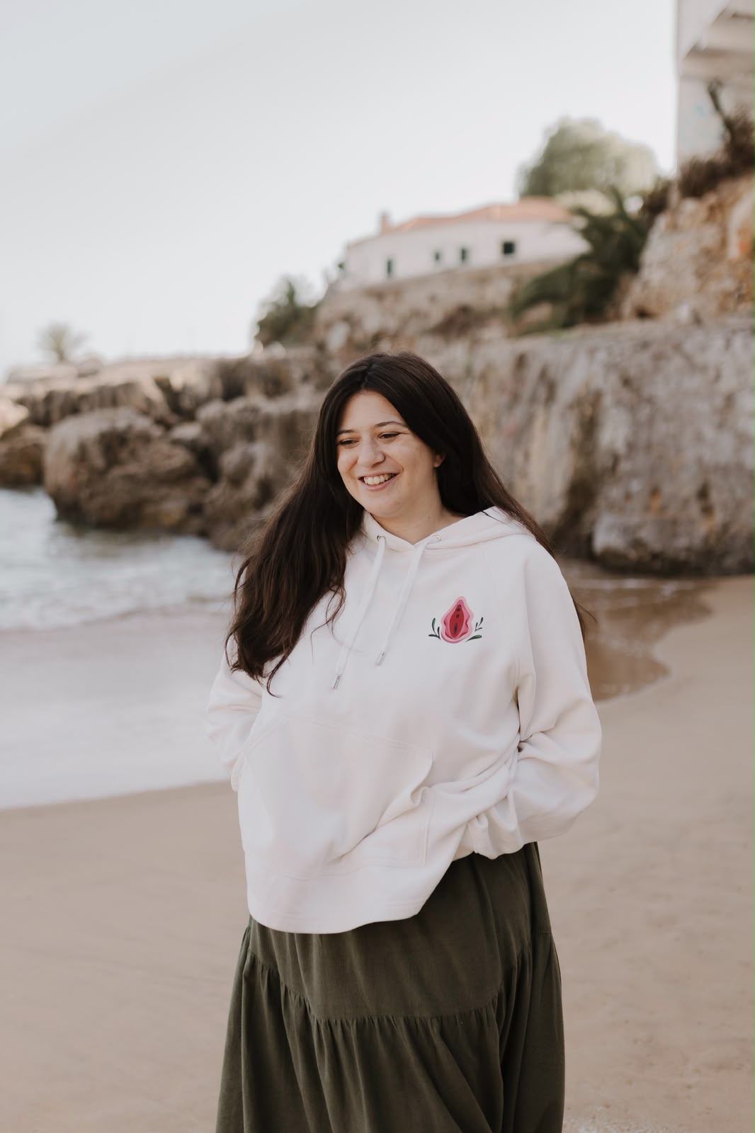 Mulher a sorrir na praia com uma hoodie creme com a ilustração de uma vulva