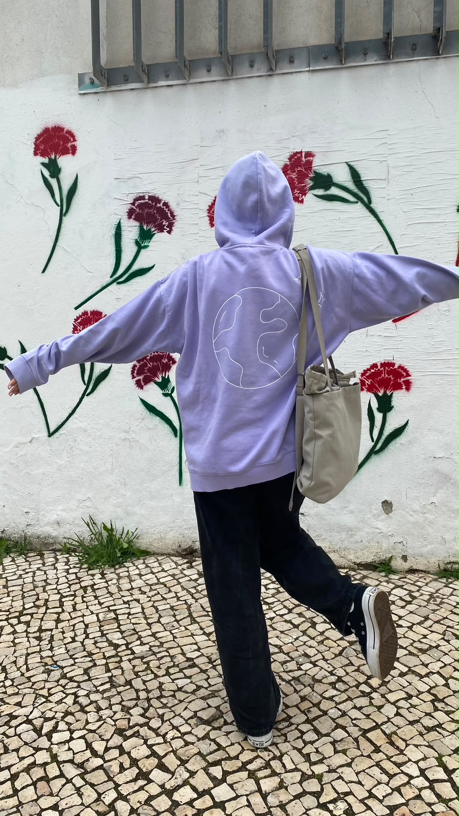 Pessoa com uma hoodie lilás com um globo desenhado, a dançar em frente a uma parede pintada com cravos vermelhos