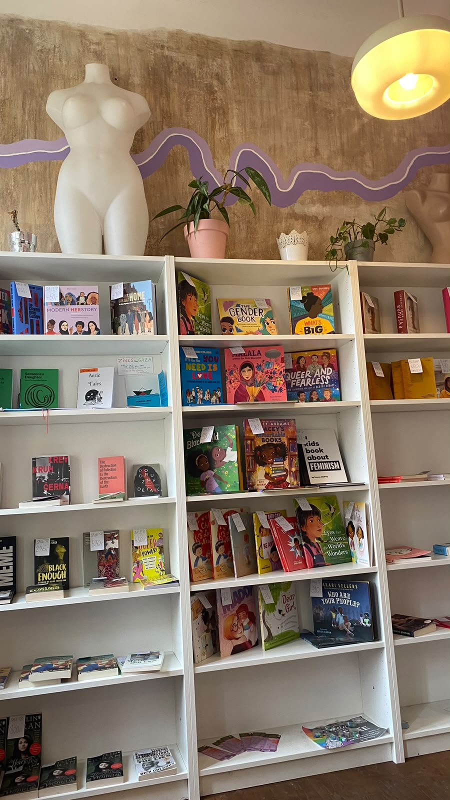 Estantes com livros feministas e queer, por baixo de uma escultura branca de um corpo feminino