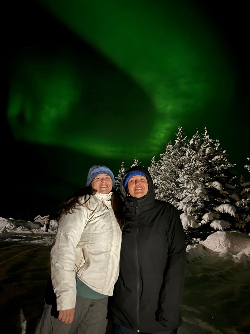Mariana e uma amiga sorridentes debaixo de uma aurora boreal verde, junto a árvores cobertas de neve