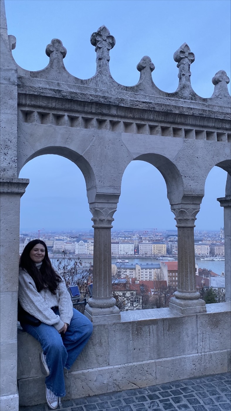 Mariana sentada nos arcos de pedra do Bastião dos Pescadores, com Budapeste ao fundo