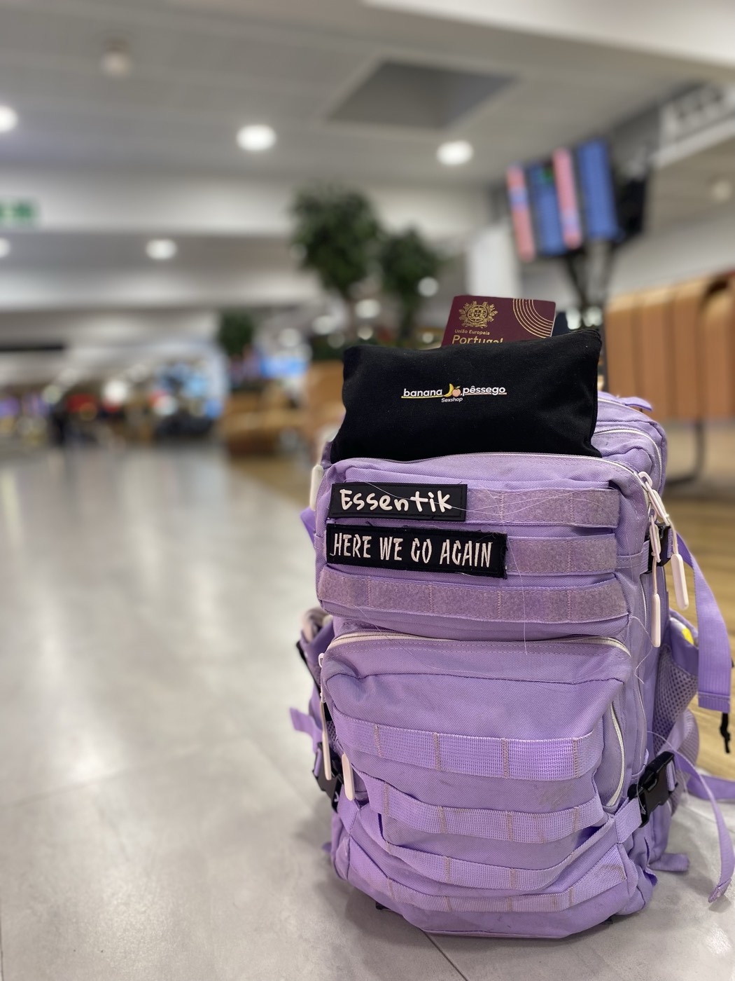 Mochila lilás com um emblema que diz Here we go again e um passaporte português em cima, num aeroporto