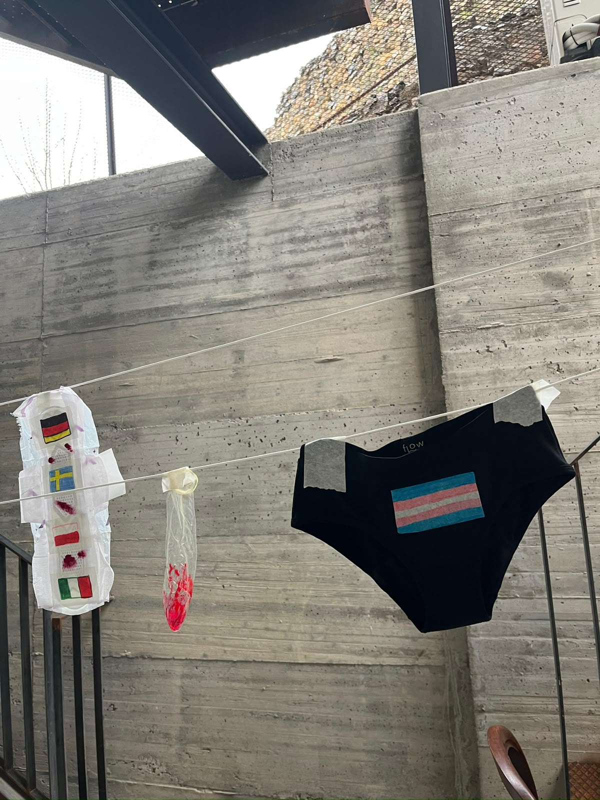 Estendal com um penso decorado com bandeiras de países, um preservativo e cuecas menstruais com a bandeira trans